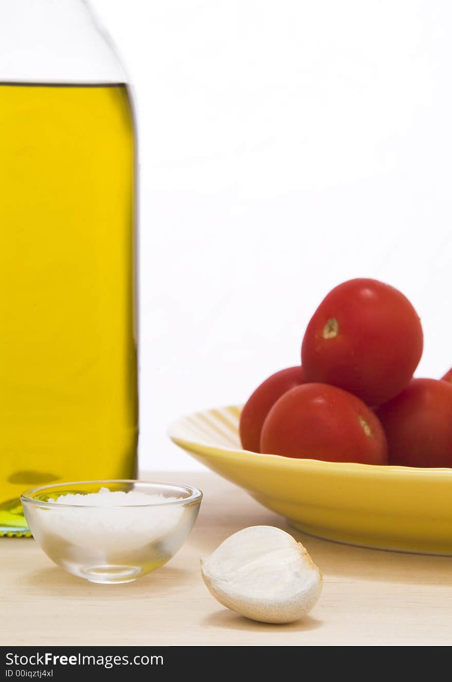 Olive oil bottle, small portion of salt, garlic clove, and a yellow plate with tomatoes on top of a wooden cutting board against a white background. Olive oil bottle, small portion of salt, garlic clove, and a yellow plate with tomatoes on top of a wooden cutting board against a white background.