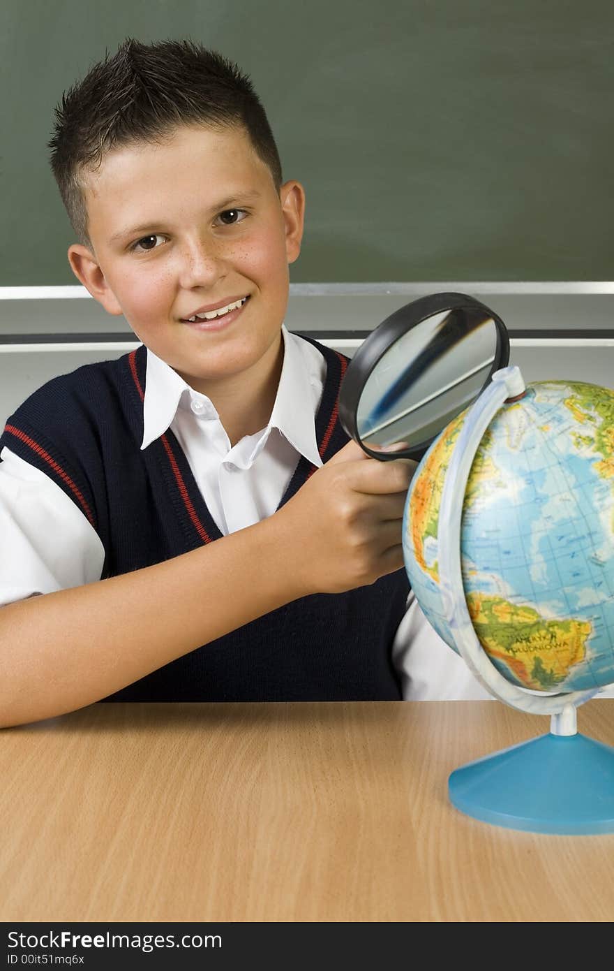 Young, smiling boy sitting at desk, nearby globe. Holding magnifying glass. Looking at camera, front view. Young, smiling boy sitting at desk, nearby globe. Holding magnifying glass. Looking at camera, front view