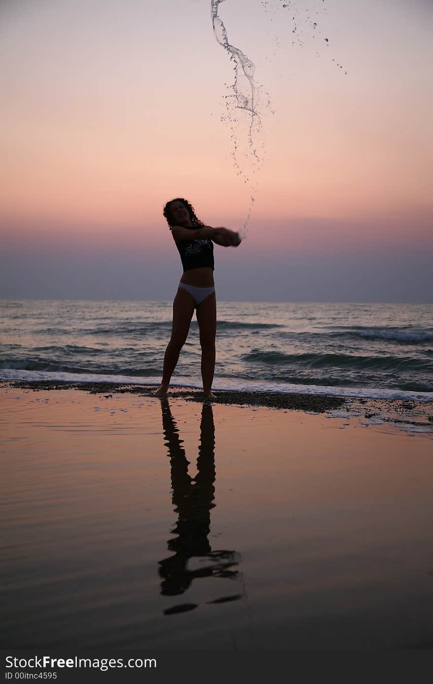 Young woman sprinkles by water on beach on sunset