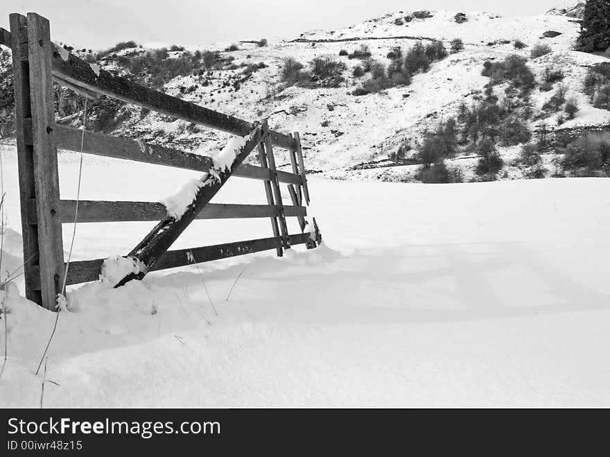 A monochrome image of a snowy gate opeing onto a snow covered field