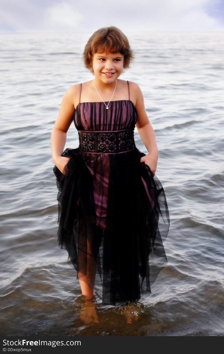 Full-body portrait of an elementary girl in formalwear holding up her skirt while standing in lake water. Full-body portrait of an elementary girl in formalwear holding up her skirt while standing in lake water.