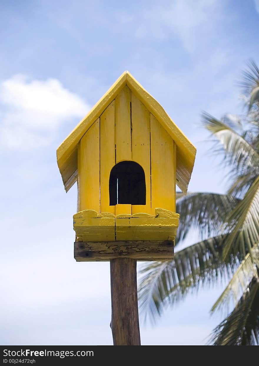 Yellow birdhouse on a background of palm trees (out of focus) and cloudy, blue sky.