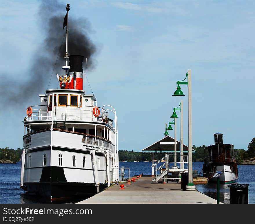 Two vintage steamships at dock.  The larger, white one is puffing dark black smoke.