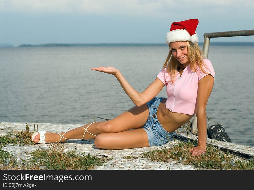 The young beautiful woman sits on seacoast with Santa Claus hat. The young beautiful woman sits on seacoast with Santa Claus hat