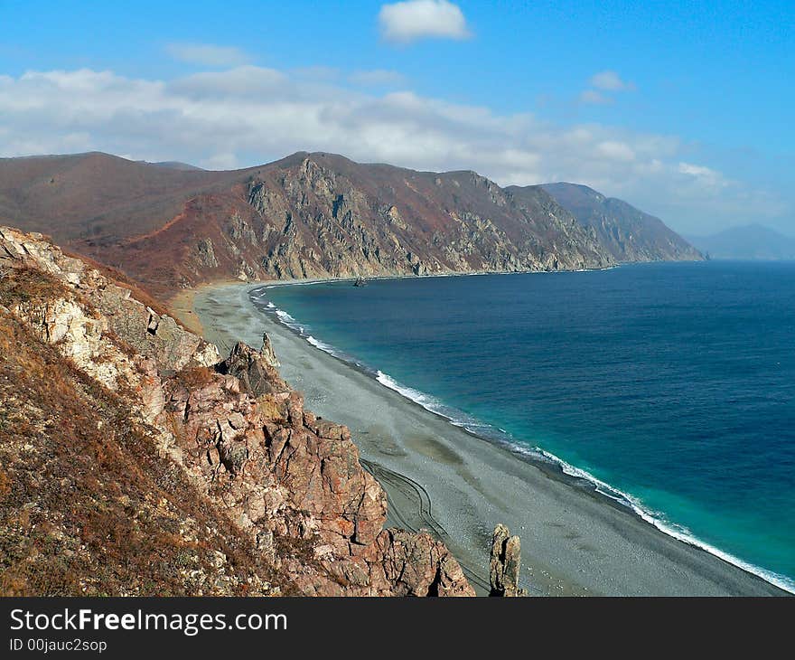 A cloudscape on sea: turquoise sea, grey beach, yellow-red capes and rocks, white surf and white clouds on azure sky. Autumn. Russian Far East, Primorye, Japanese sea, Tasovaya bay. A cloudscape on sea: turquoise sea, grey beach, yellow-red capes and rocks, white surf and white clouds on azure sky. Autumn. Russian Far East, Primorye, Japanese sea, Tasovaya bay.