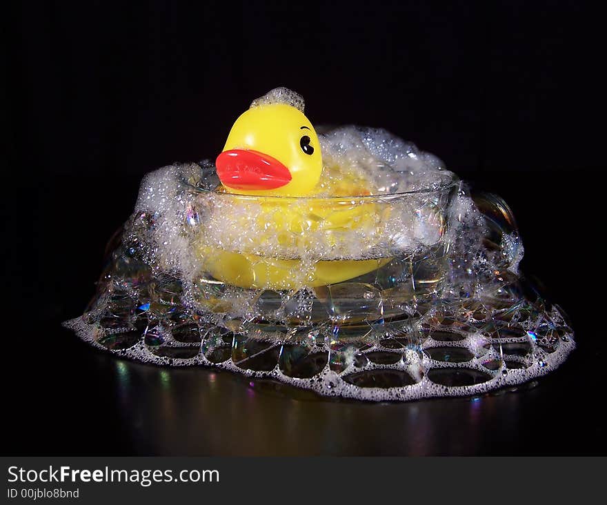 Bright yellow rubber duck taking a bubble bath in a small, clear glass dish with bubbles falling all around the outside of the dish.  This photo has a black background. Bright yellow rubber duck taking a bubble bath in a small, clear glass dish with bubbles falling all around the outside of the dish.  This photo has a black background.