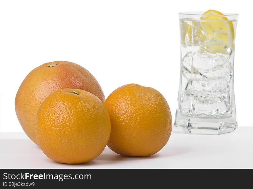 Two oranges and a grapefruit with a glass of sparkling water in the back filled with ice and a slice of lemon. Two oranges and a grapefruit with a glass of sparkling water in the back filled with ice and a slice of lemon