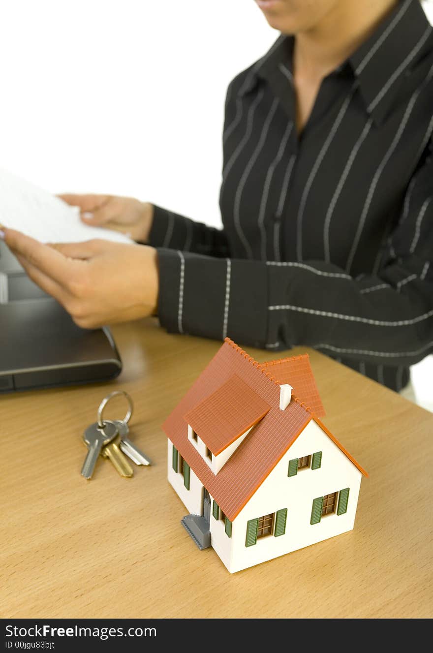 Young woman sitting at desk in front of laptop, keys and house miniature. Holding piece of paper. White background, side view. Young woman sitting at desk in front of laptop, keys and house miniature. Holding piece of paper. White background, side view