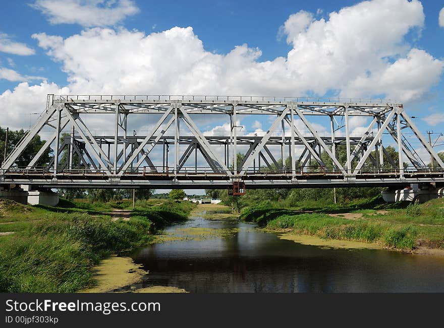 Grey railway bridge over small river with green banks and the cloudy summer sky