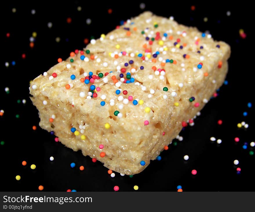 Close up color photograph of a delicious marshmallow crispy dessert bar with colorful sprinkles on and around it.  Black background.