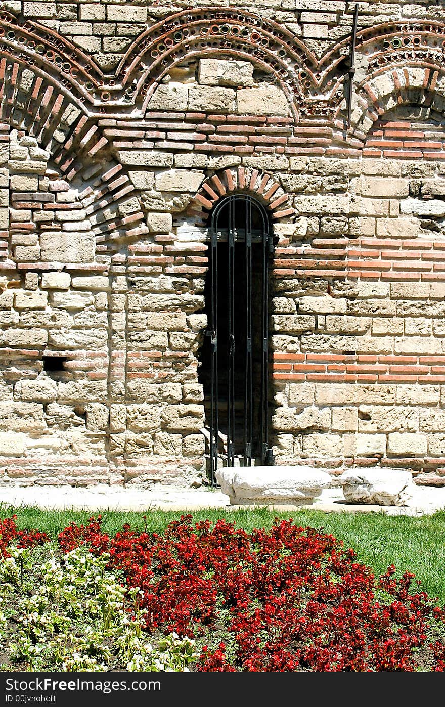 An ancient wall with the entrance with the grating and little flowerbed ahead. An ancient wall with the entrance with the grating and little flowerbed ahead