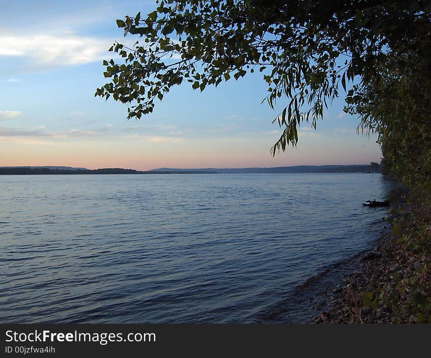 Branch above the water. Blue-rose sky. Little wave on the water.