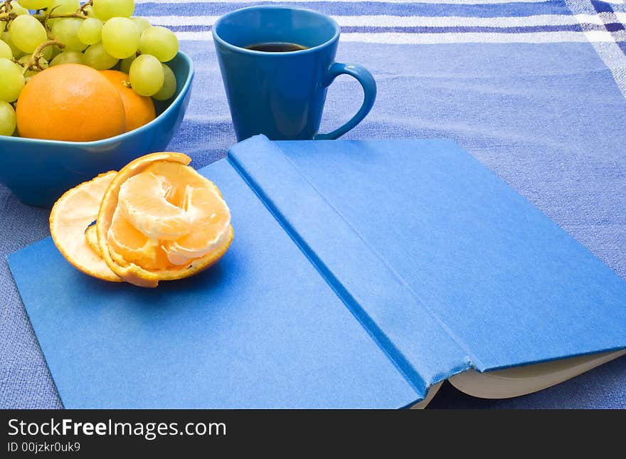 A book and a cup of coffee and some fruit on a blue tablecloth. A book and a cup of coffee and some fruit on a blue tablecloth.