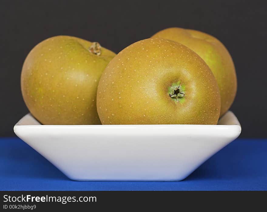 A white bowl containing three golden Russet apples, against a black and blue background. A white bowl containing three golden Russet apples, against a black and blue background.