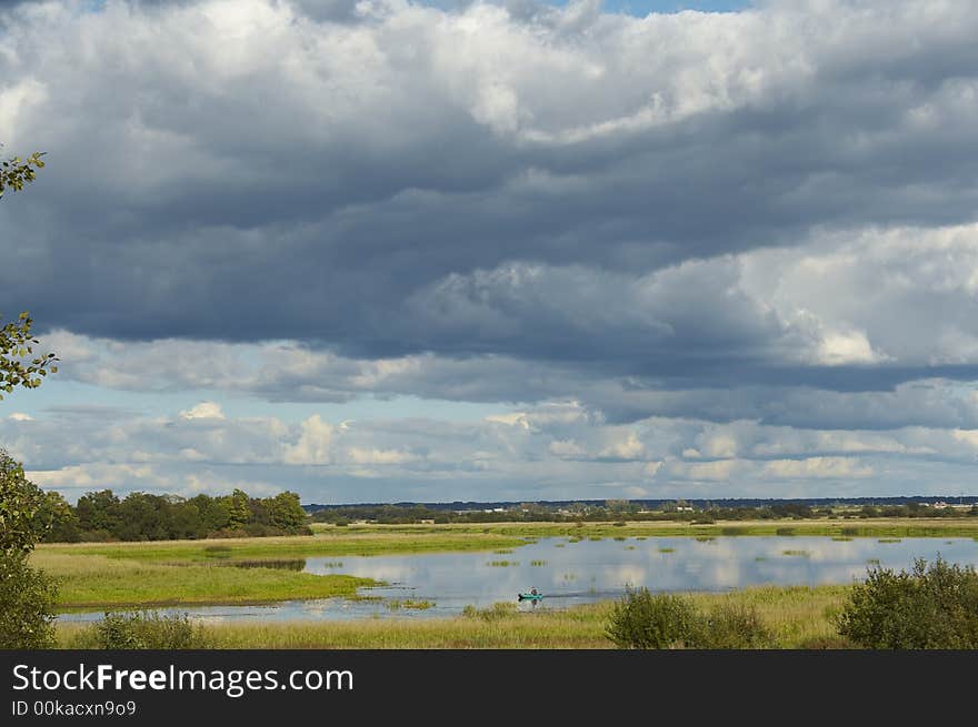 White fluffy clouds, blue sky and green wood reflecting in a clear, placid lake.