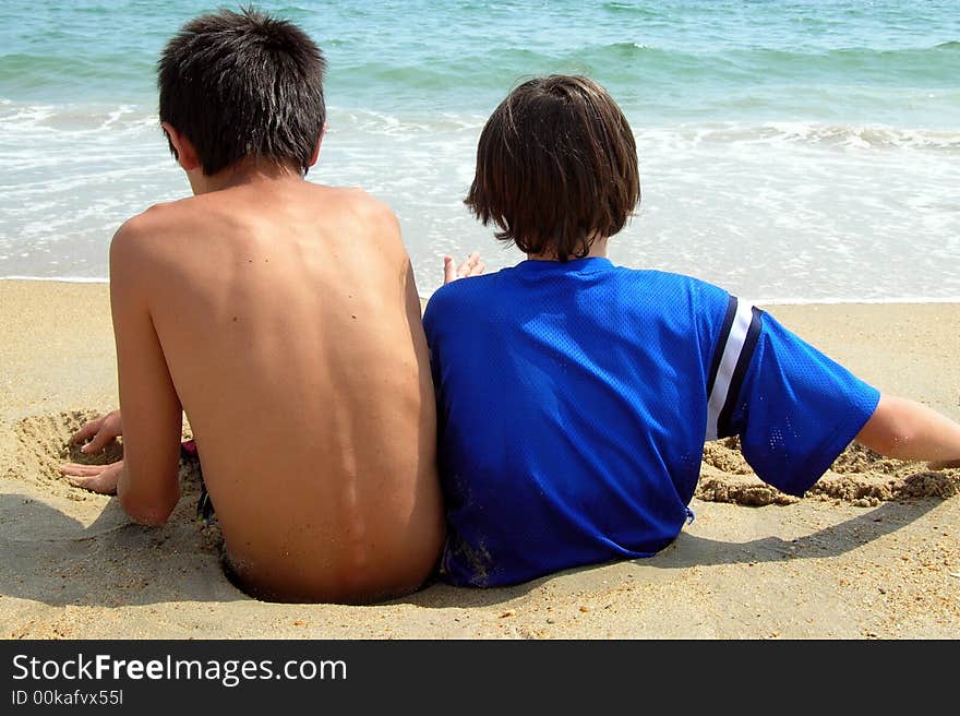 2 boys sit deep in the sand facing the ocean, letting the waves dig them in deeper and deeper. 2 boys sit deep in the sand facing the ocean, letting the waves dig them in deeper and deeper.