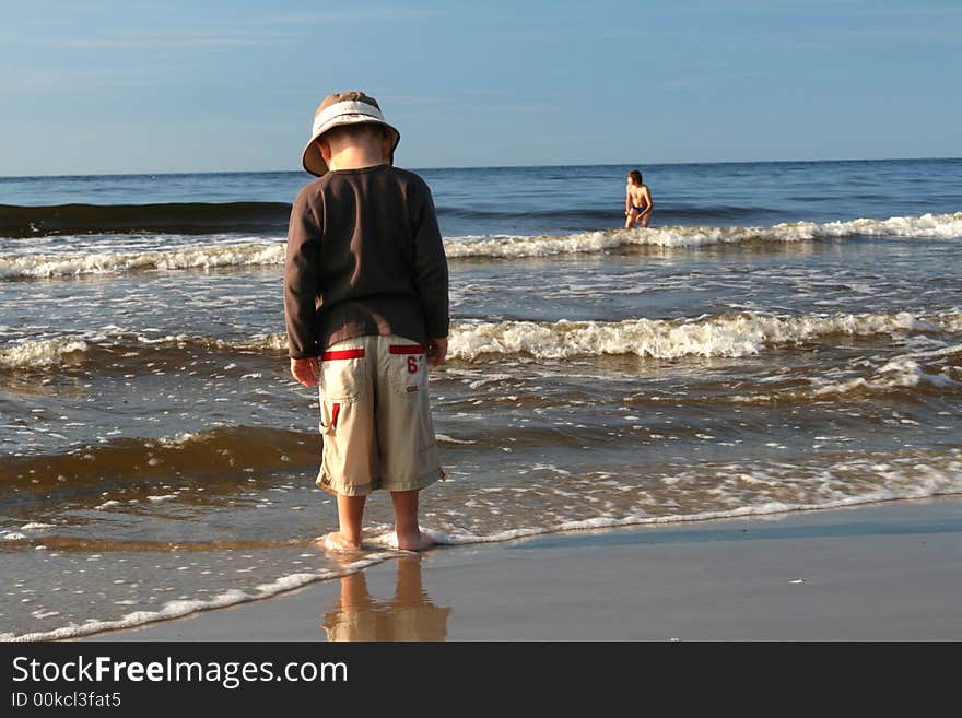 3 years old boy standing on the shore of a sandy beach and looking out over the sea as waves crash at his feet. 3 years old boy standing on the shore of a sandy beach and looking out over the sea as waves crash at his feet.