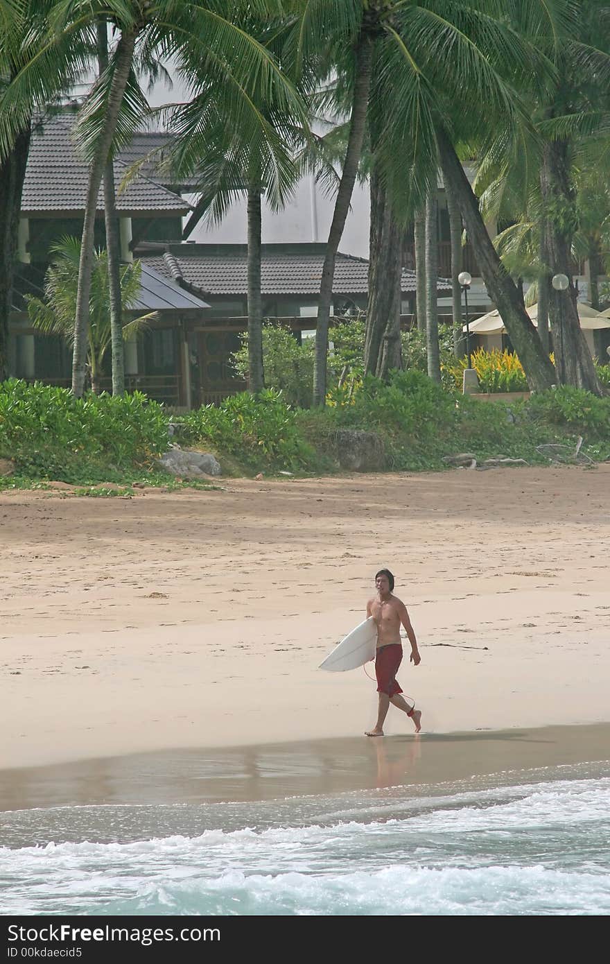 A surfer walks along the waters edge on a tropical beach in Thailand. A surfer walks along the waters edge on a tropical beach in Thailand