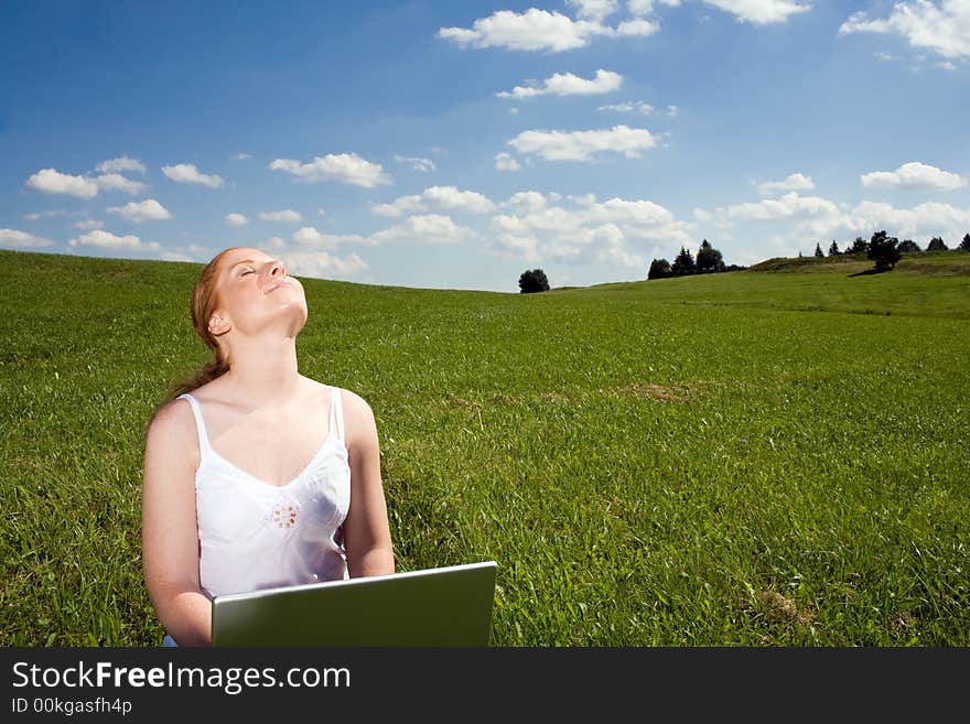 Woman sitting with laptop on a meadow going to the horizon. Blue sky with white clouds. Face towards the sun, eyes closed. Woman sitting with laptop on a meadow going to the horizon. Blue sky with white clouds. Face towards the sun, eyes closed.