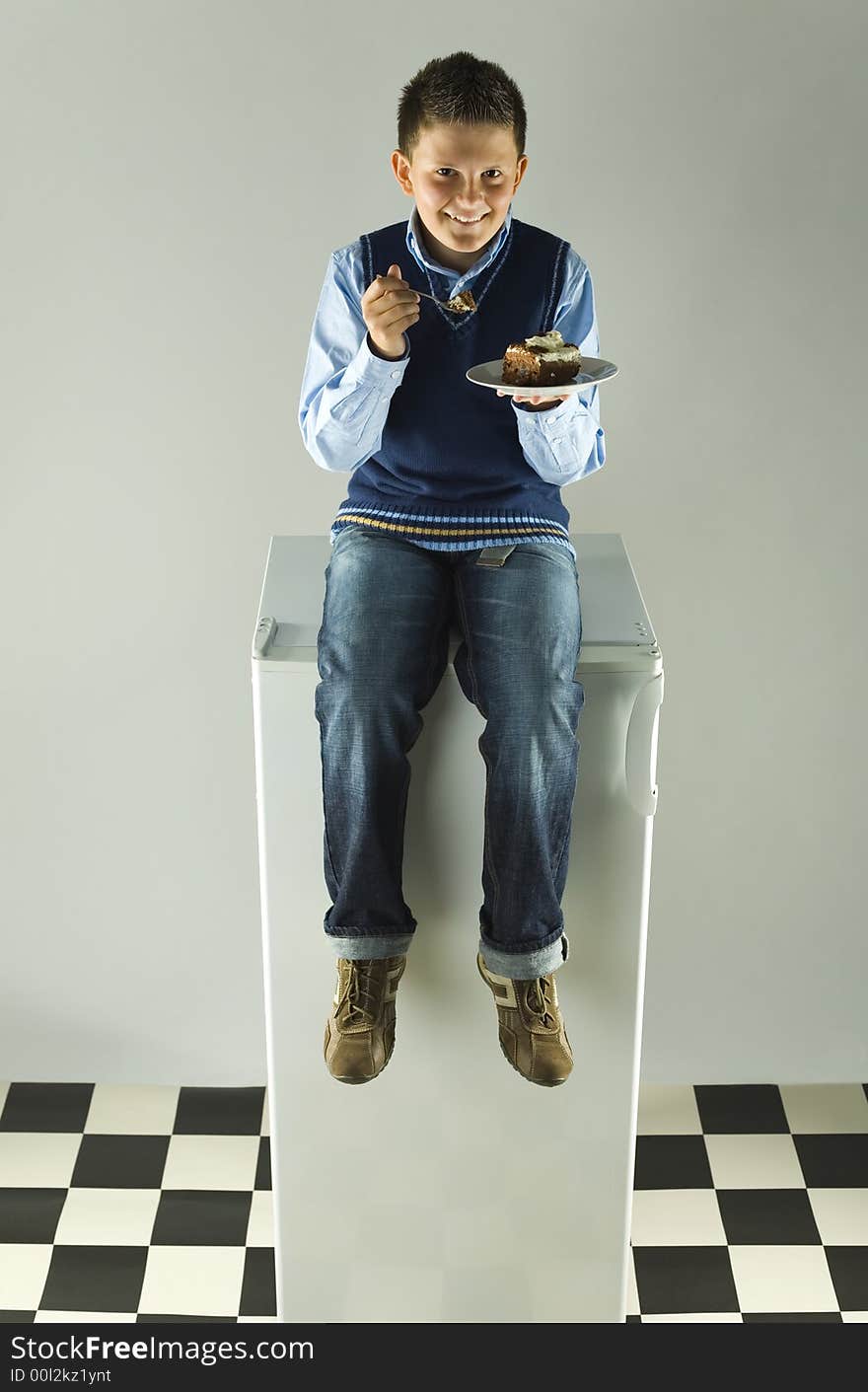 Happy boy sitting on fridge and eating cake. Front view. Grey background. Happy boy sitting on fridge and eating cake. Front view. Grey background.