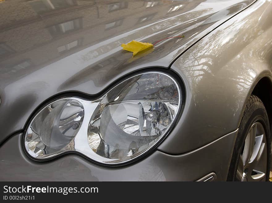 Headlight of the new machine. Metallic paint. A small yellow leaf of a maple. House and maple reflection.