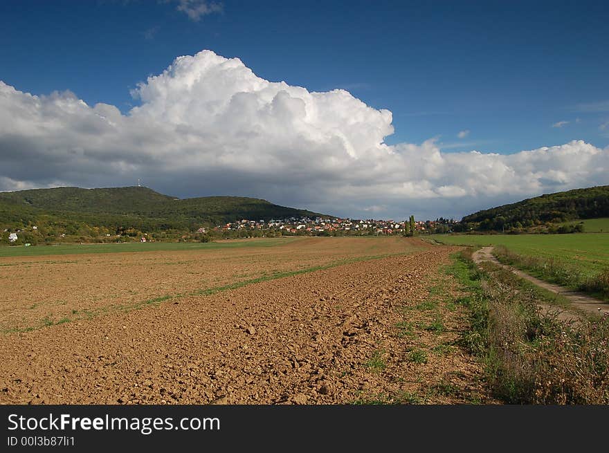 Heavy low white cloud above roofs of village with fields and lane in foreground. Heavy low white cloud above roofs of village with fields and lane in foreground