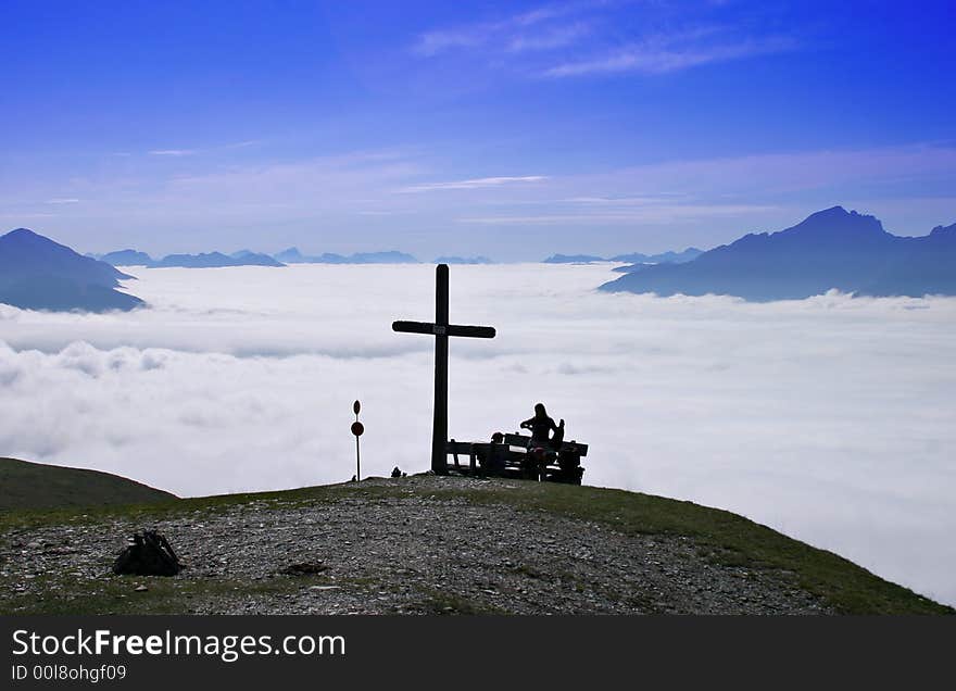 Hikers sitting on a bench close to a summit cross, high above the cloud cover stretching to the background with distant mountain peaks. Hikers sitting on a bench close to a summit cross, high above the cloud cover stretching to the background with distant mountain peaks