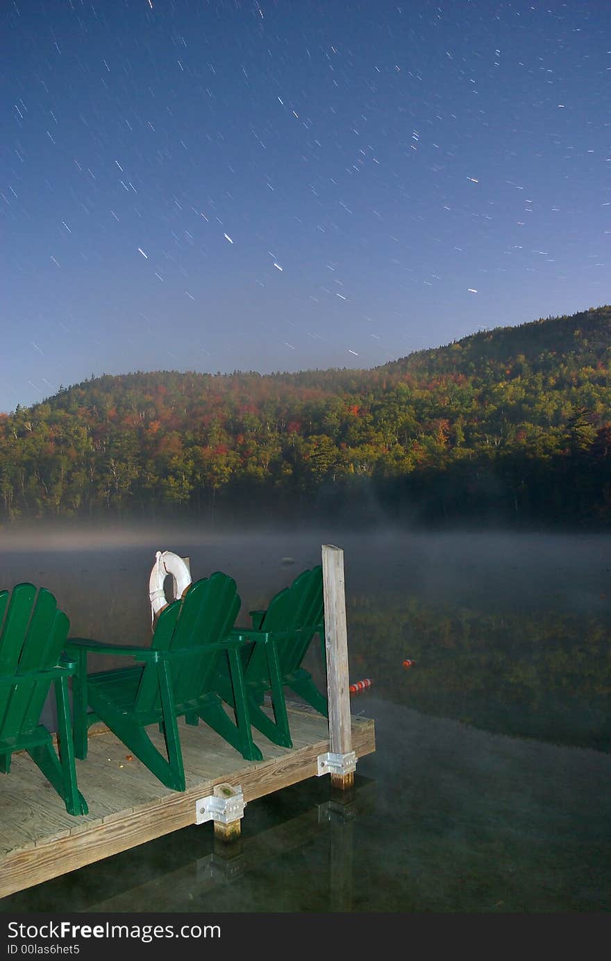 Star trails and fog late at night over a lake in the Adirondack Mountains.  Empty lawn chairs look out at the Autumn foliage. Star trails and fog late at night over a lake in the Adirondack Mountains.  Empty lawn chairs look out at the Autumn foliage.