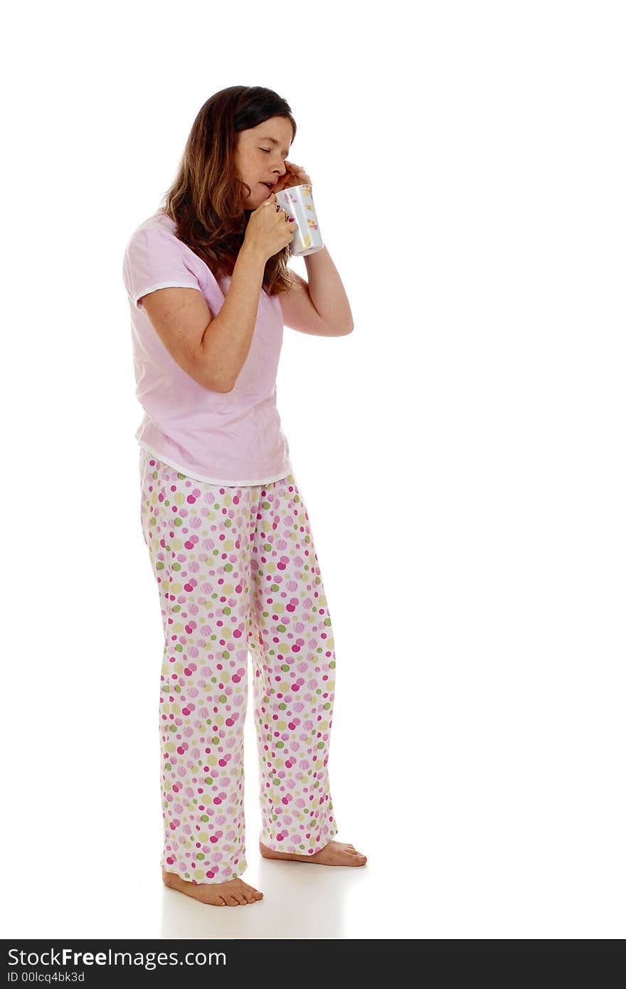 Full-length portrait of a woman in her pajamas rubbing her closed eyes while trying to drink her morning coffee.