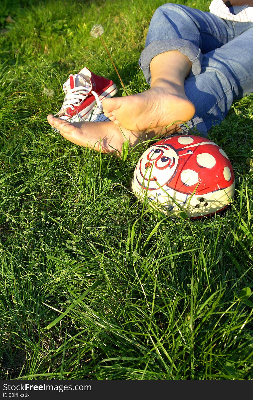 View of girl's legs and her bare feet on grass next to pair of athletic shoes. Face of girl not in photo. View of girl's legs and her bare feet on grass next to pair of athletic shoes. Face of girl not in photo.