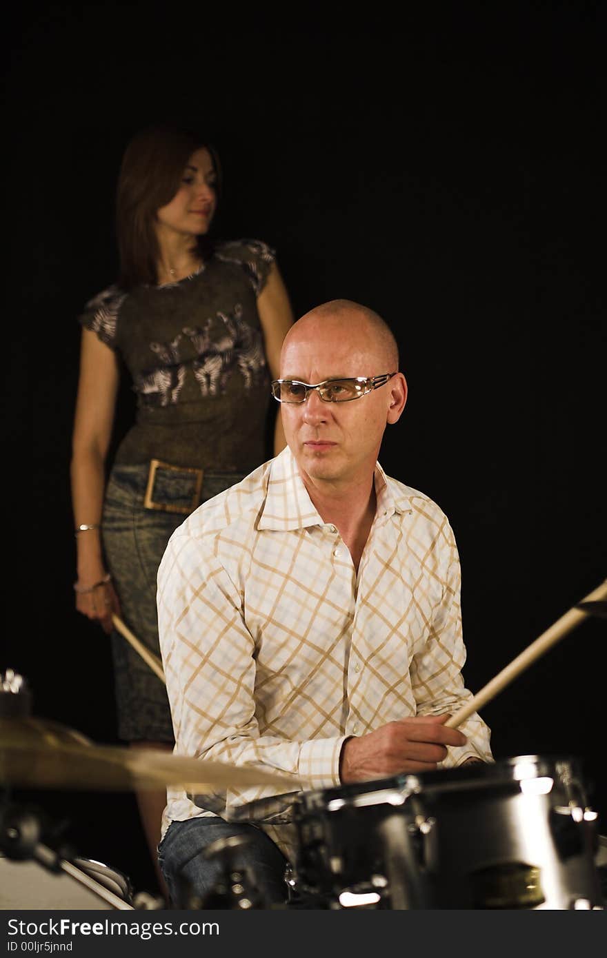 Drummer playing over black backdrop with woman standing in background