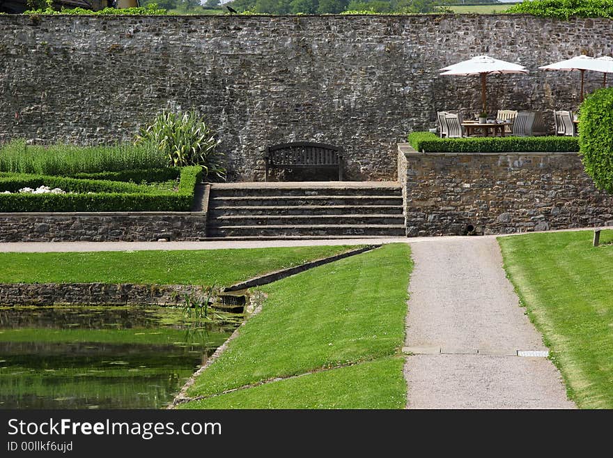 Ancient walled garden in summer, with outdoor patio dining area with tables, chairs and sun umbrellas, a bench, areas of lawn, shrubs and flowers and a pond.