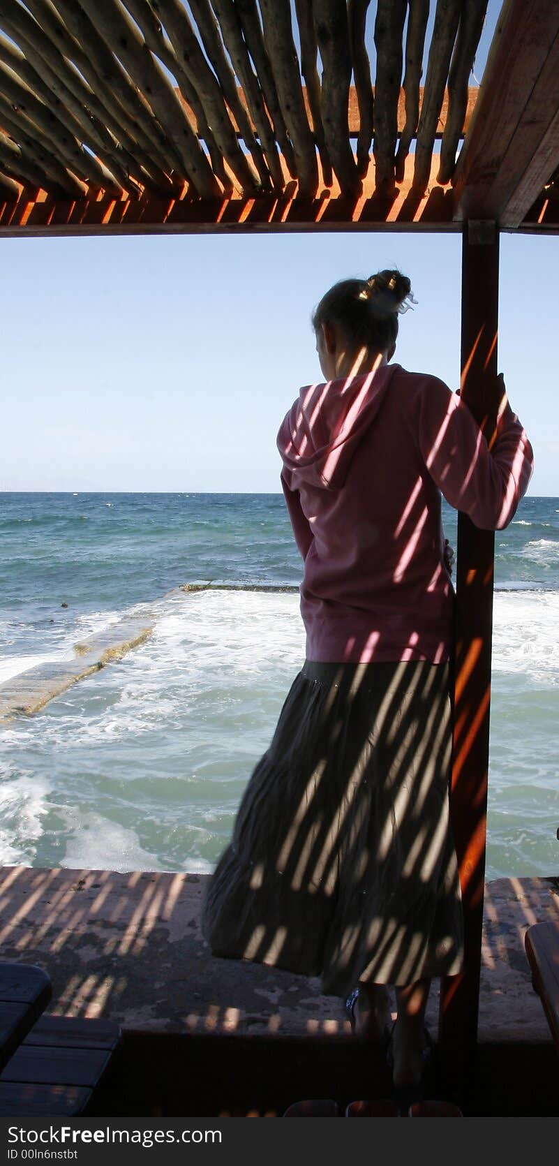 Young girl deep in thought while looking out at the sea. Tidal pool in foreground. Striped effect from sunlight through pole ceiling. Young girl deep in thought while looking out at the sea. Tidal pool in foreground. Striped effect from sunlight through pole ceiling.