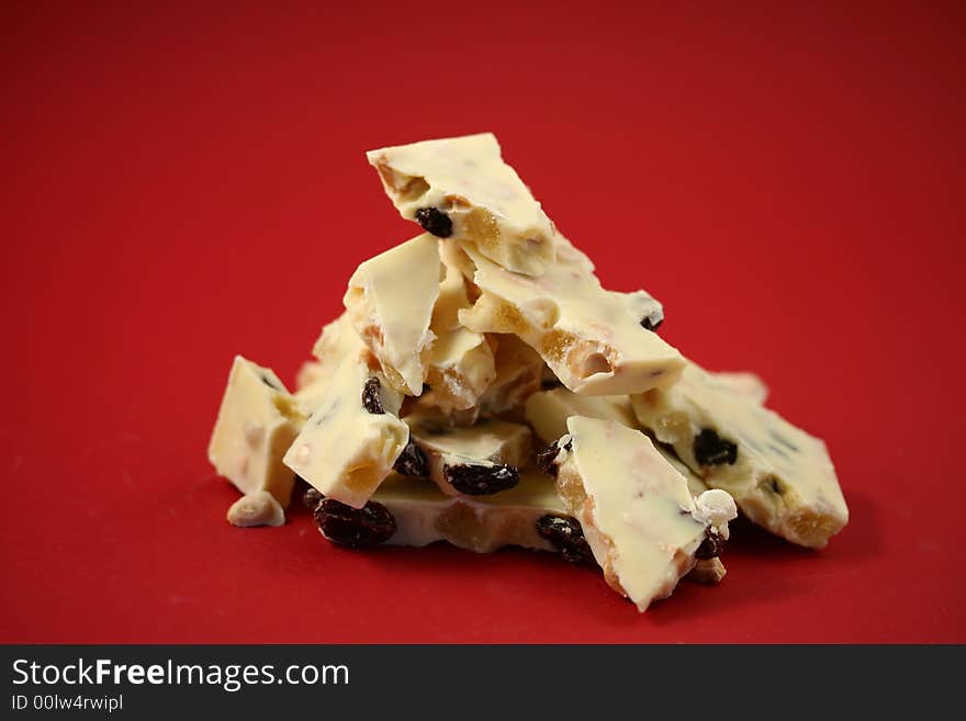 Detail of a pile of pieces of a white chocolate slab with jelly, raisins and peanuts, red background. Detail of a pile of pieces of a white chocolate slab with jelly, raisins and peanuts, red background.