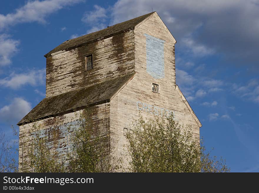 An old grain elevator, with peeling paint, surrounded by leafy trees and backed by a blue sky and white clouds.