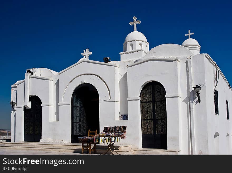 A white Greek orthodox church on a clear blue sky background with a trinket stall.