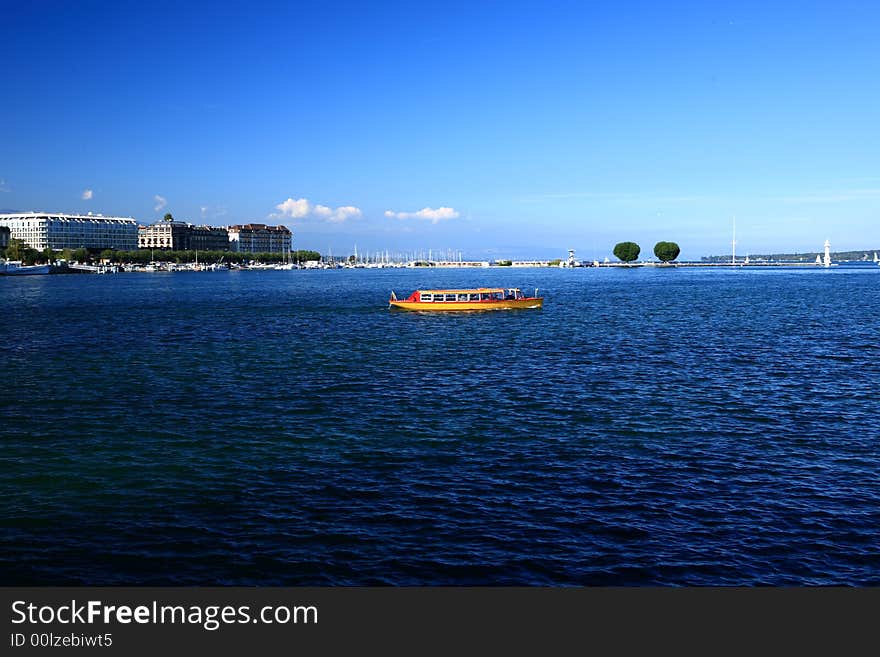 The yellow boat in Leman Lake, and the blue water, blue sky and white clouds, and the buildings and trees at the far end of the lake, forms a peaceful senery.