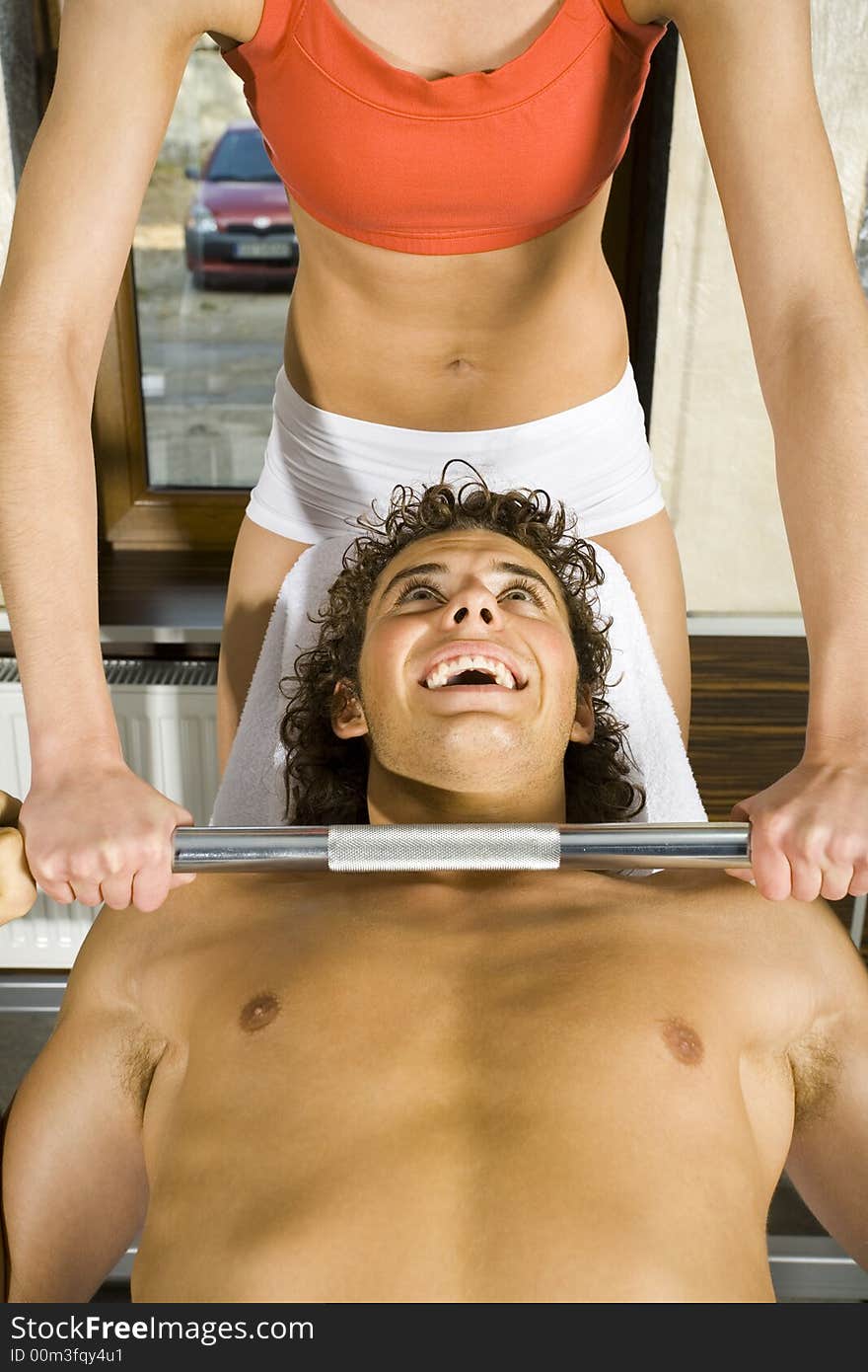 Young couple, working out in gym. Woman is helping man. Man is lying on bench and picking up dumbbell. Smiling and looking at woman. Front view. Young couple, working out in gym. Woman is helping man. Man is lying on bench and picking up dumbbell. Smiling and looking at woman. Front view