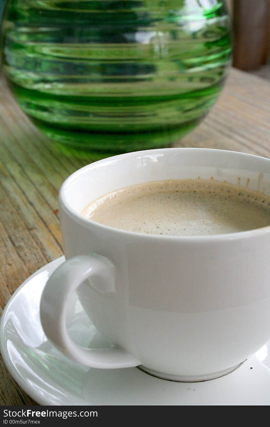 Close-up of a white coffee cup on a timber table with a green glass vase in the background. Close-up of a white coffee cup on a timber table with a green glass vase in the background