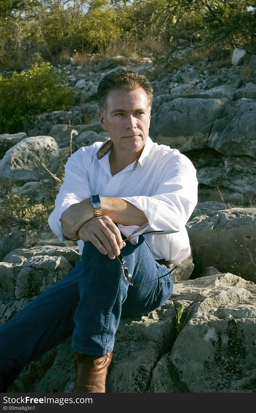 A man, sitting leisurely on a boulder on the side of a rocky hill. A man, sitting leisurely on a boulder on the side of a rocky hill.