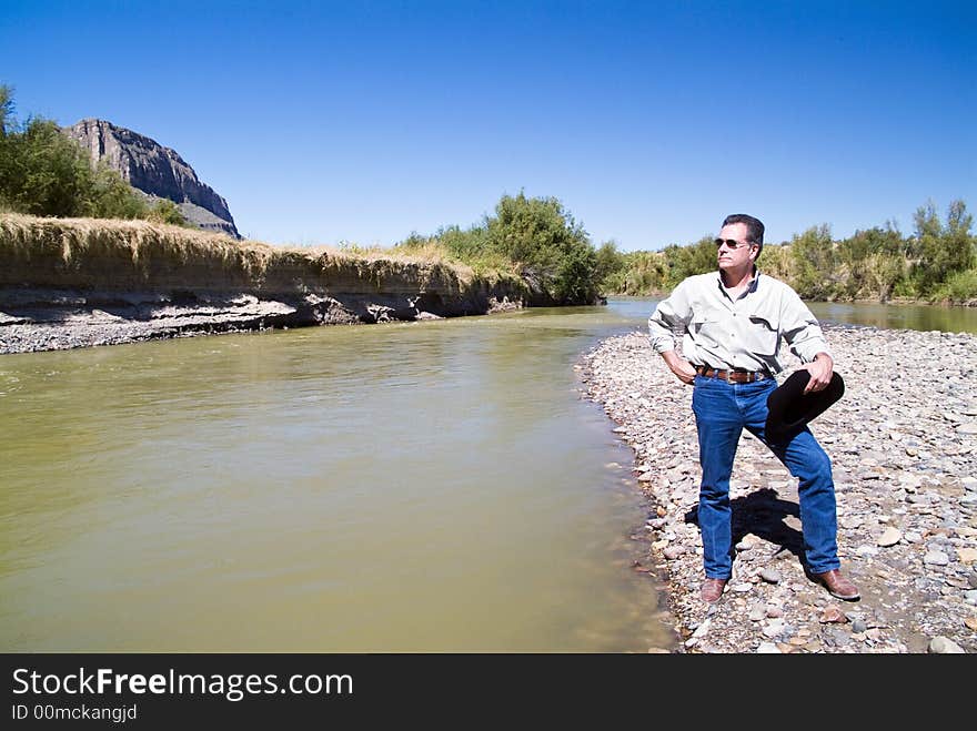 A man, hat in hand, standing beside a shallow portion of a river looking up at the cliffs. A man, hat in hand, standing beside a shallow portion of a river looking up at the cliffs.