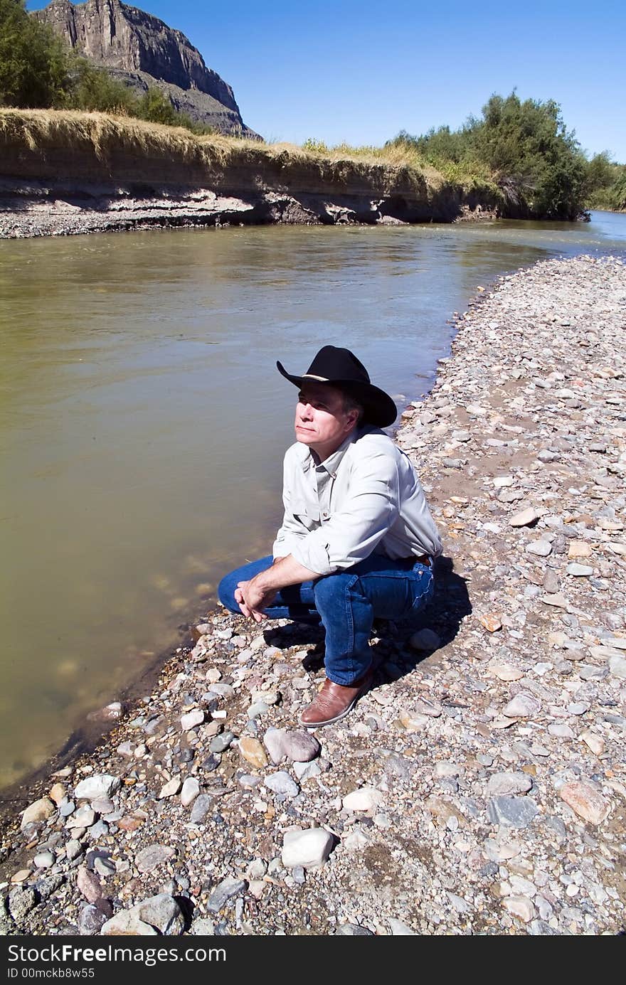 A man dressed in western garb squatting beside a river with an appreciative expression on his face. A man dressed in western garb squatting beside a river with an appreciative expression on his face.