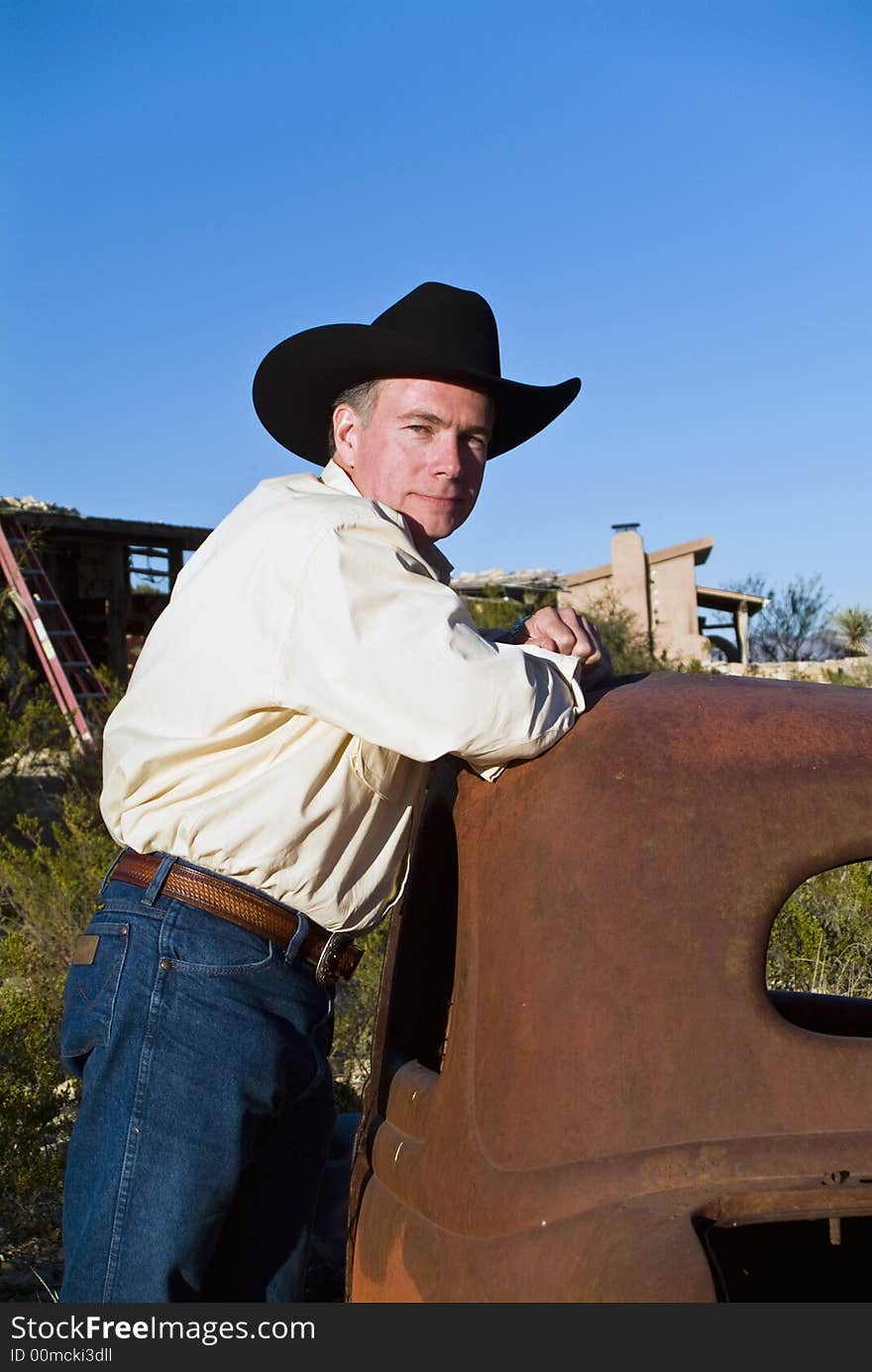 A friendly looking man in a black cowboy hat leaning on an old, long abandoned car. A friendly looking man in a black cowboy hat leaning on an old, long abandoned car.