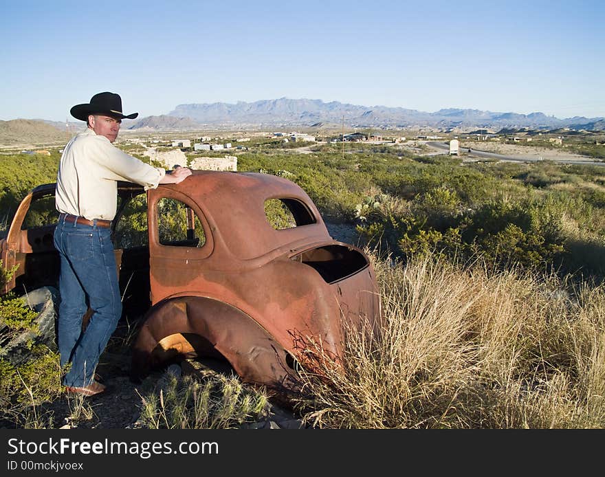 A man in western attire leaning on an old, long abandoned vehicle surrounded by rugged terrain.