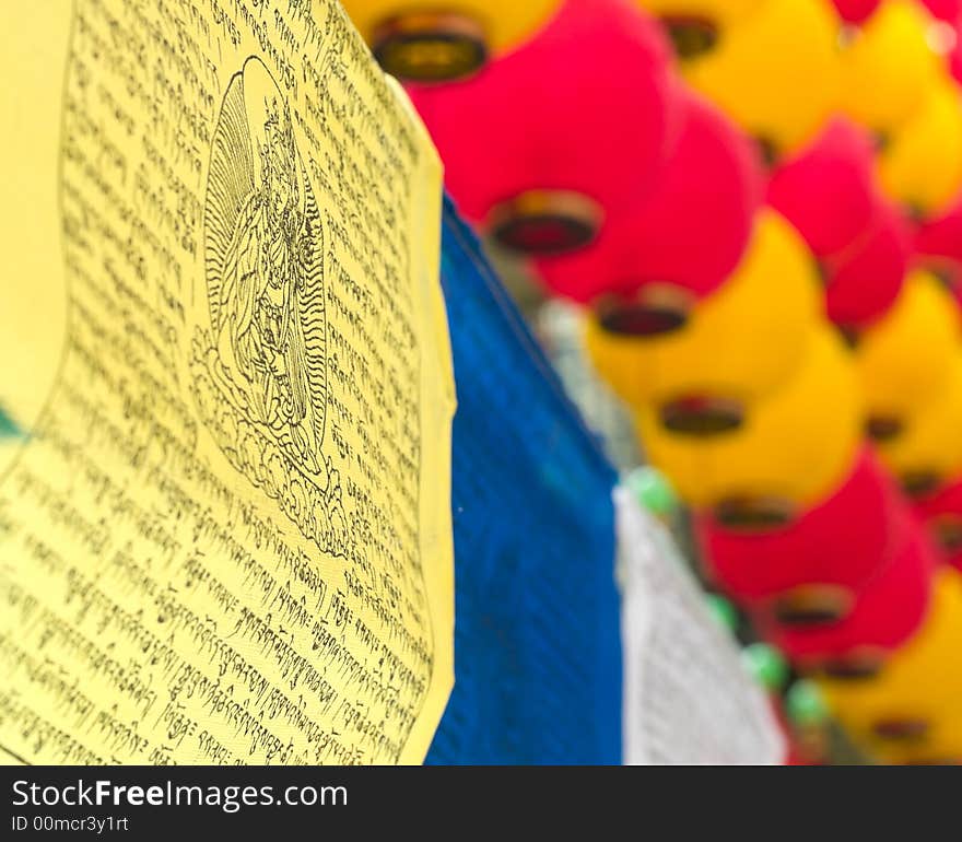 A row of prayer colored prayer flags with mantras inscribed in Sanskrit stringed below red and yellow lanterns. A row of prayer colored prayer flags with mantras inscribed in Sanskrit stringed below red and yellow lanterns