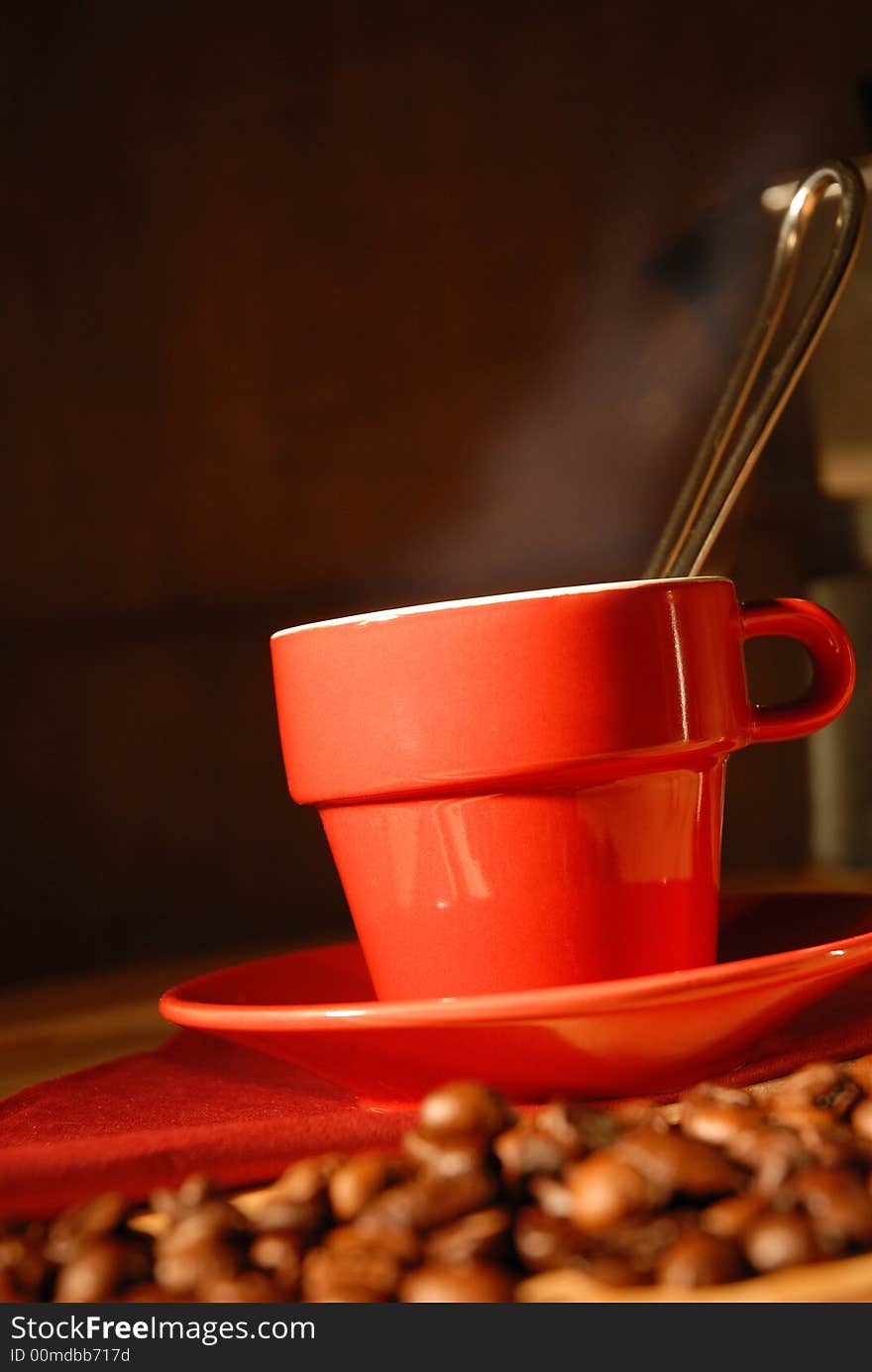 Warm atmosphere for a close-up of a red cup of coffee. Out of focus a moka on the background and coffee beans in first plan to complete the composition. Warm atmosphere for a close-up of a red cup of coffee. Out of focus a moka on the background and coffee beans in first plan to complete the composition.