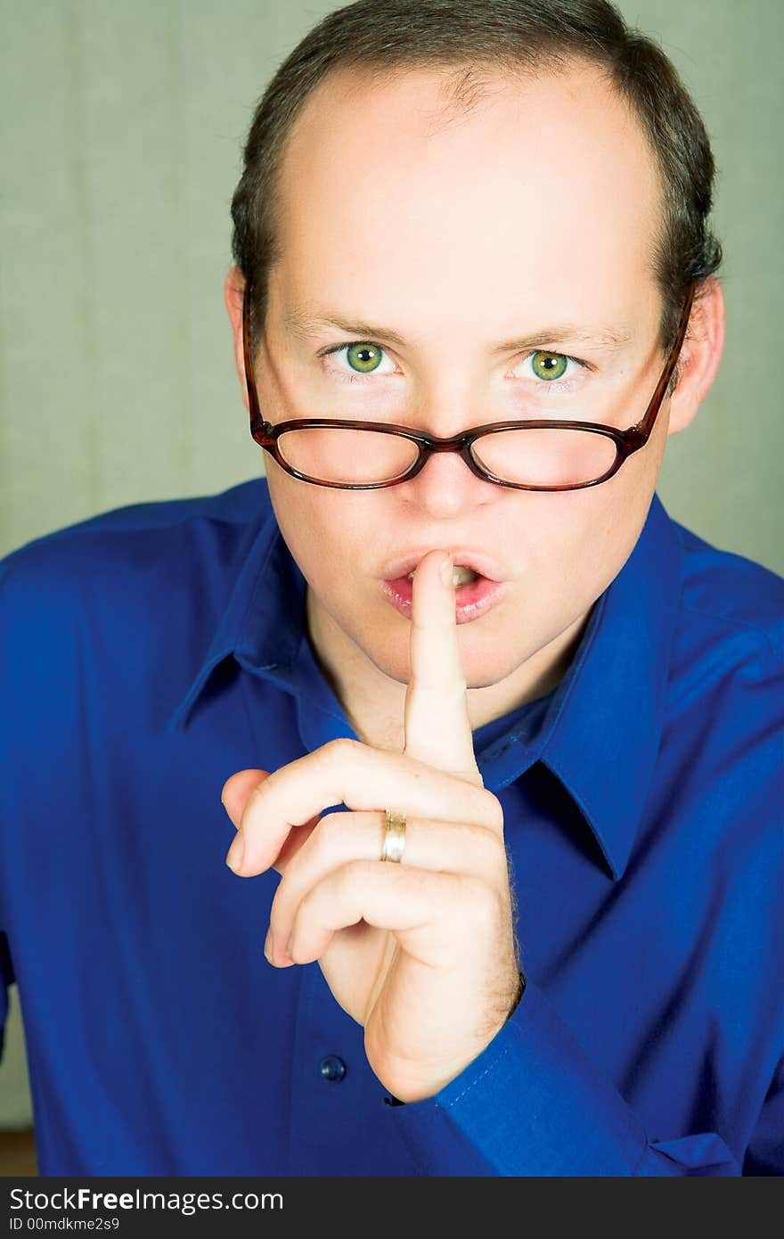 Man with green eyes in blue shirt wearing glasses demanding silence with finger next to his mouth. Man with green eyes in blue shirt wearing glasses demanding silence with finger next to his mouth