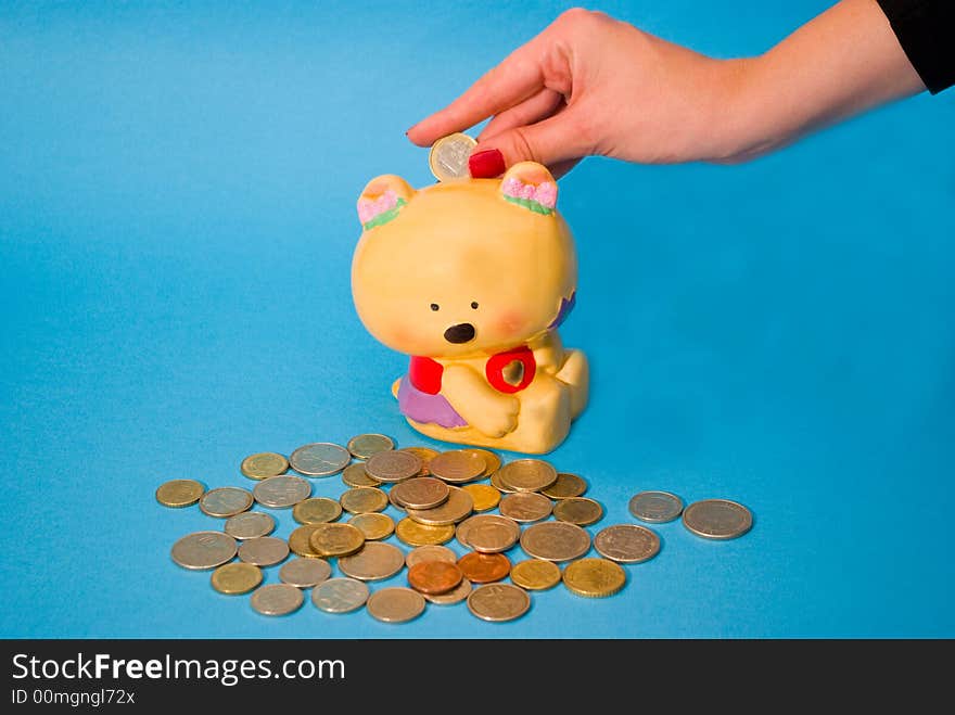 A girl's hand, putting a coin inside a toy safe, with a bunch of coins scattered on a blue background. A girl's hand, putting a coin inside a toy safe, with a bunch of coins scattered on a blue background