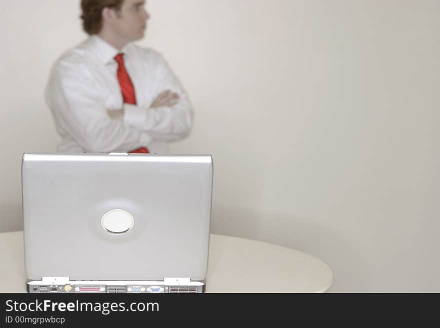 One young businessman standing with arms crossed wearing white shirt and red tie with laptop in foreground resting on white table. One young businessman standing with arms crossed wearing white shirt and red tie with laptop in foreground resting on white table