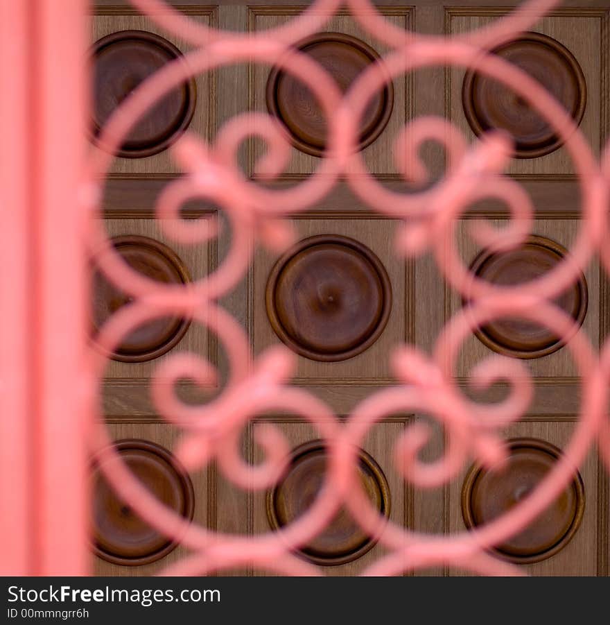 Wooden door decorated with square panels with an embossed circle seen behind a decorative vermilion coloured wrought iron gate. Wooden door decorated with square panels with an embossed circle seen behind a decorative vermilion coloured wrought iron gate